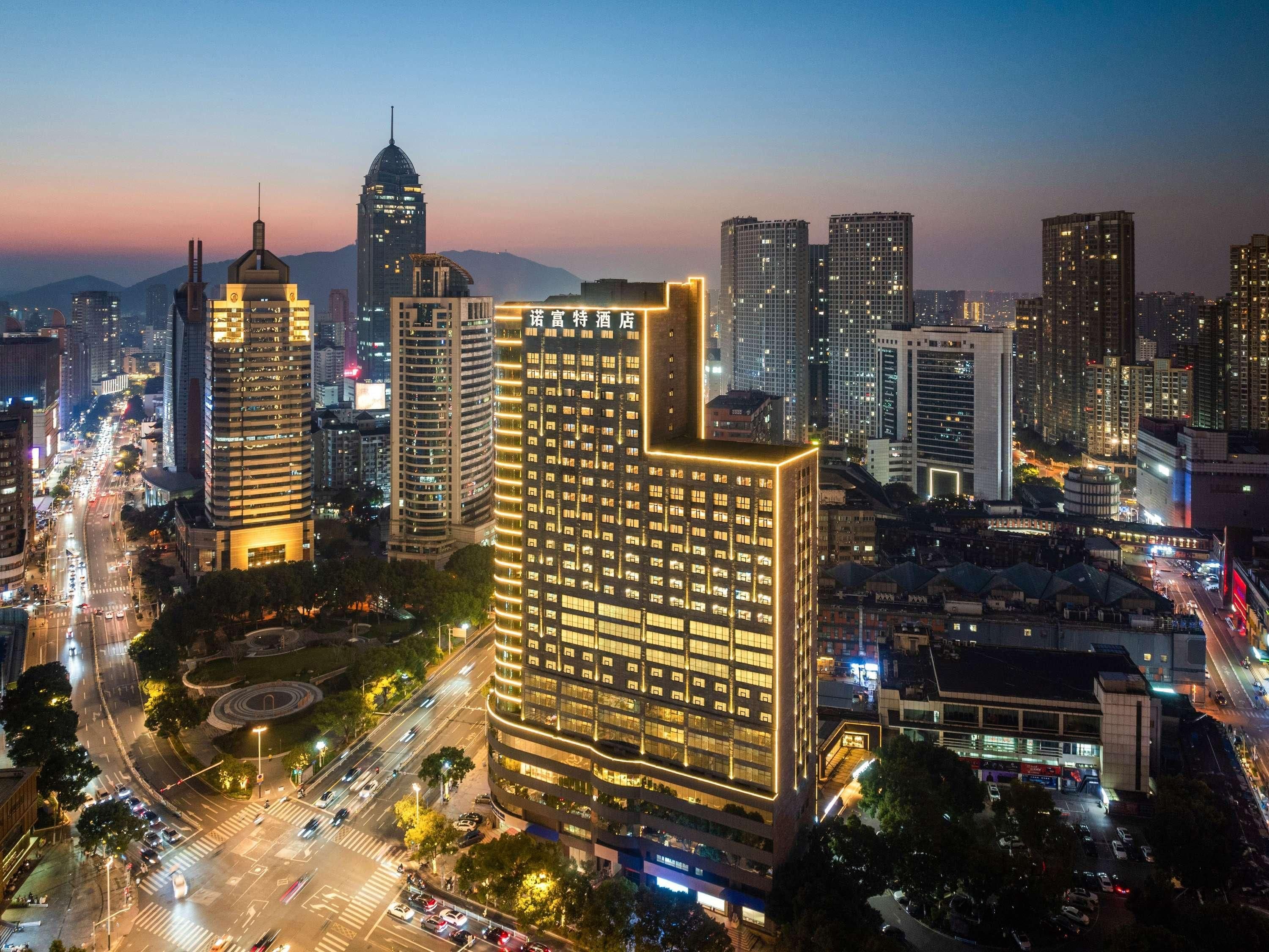 A cityscape at dusk with illuminated buildings, a prominent hotel with a distinctive corner design, and a busy intersection.