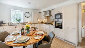 A modern kitchen with white cabinets, a wooden dining table set for four, and a clock on the wall. - 2 bedroom accommodation in Casterton, near Kirkby Lonsdale (Casterton, near Kirkby Lonsdale)