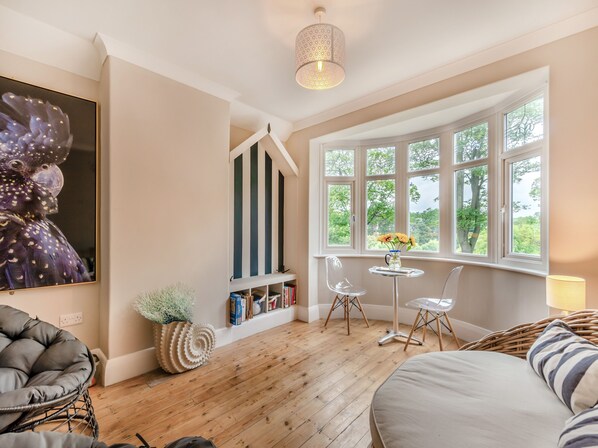 A room with a wooden floor, a large window with a view of greenery, a bookshelf, and a chair.