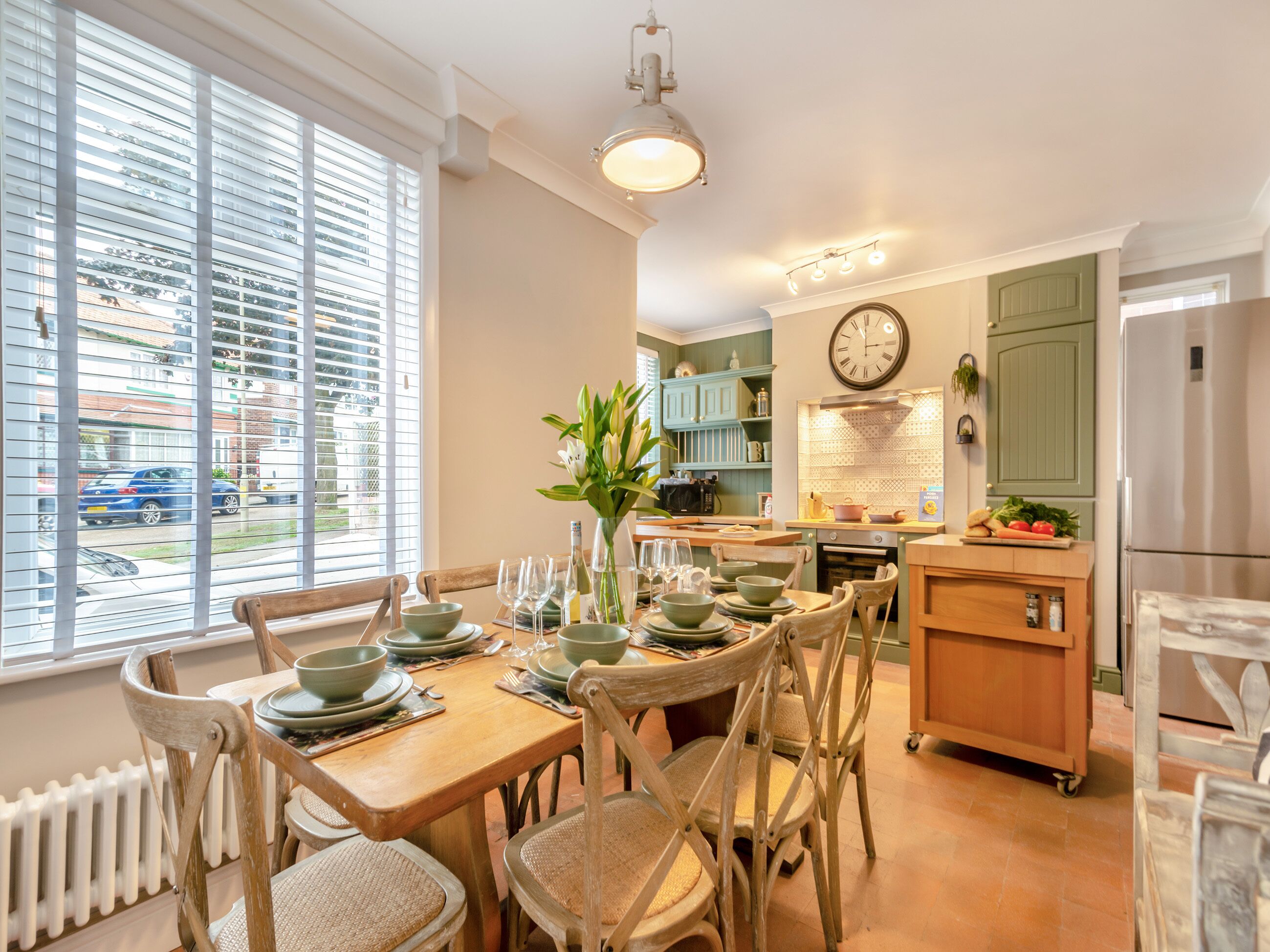 Een eetruimte met een houten tafel gedekt voor vier personen, omringd door rieten stoelen. In de achtergrond is een keuken met groene kastjes en een klok te zien.