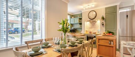 Uma sala de jantar com uma mesa de madeira posta para quatro pessoas, rodeada por cadeiras de vime. Uma cozinha com armários verdes e um relógio é visível ao fundo.