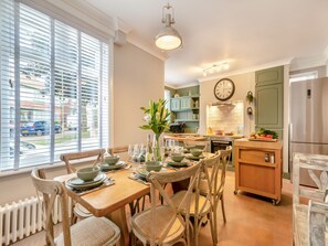 A dining area with a wooden table set for four, surrounded by wicker chairs. A kitchen with green cabinets and a clock is visible in the background.