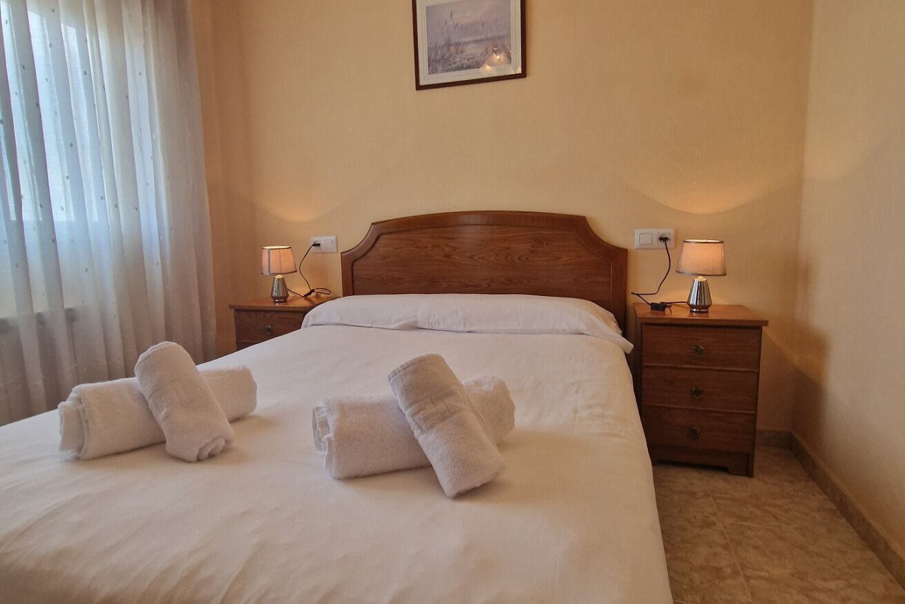 Un lit bien fait, avec du linge de lit blanc et deux serviettes enroulées, une tête de lit en bois, des lampes de chevet et un tableau encadré au mur.