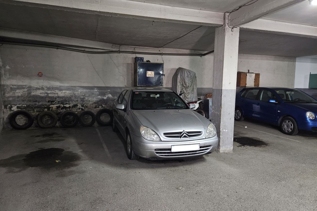 Een parkeergarage met een zilveren auto op een parkeerplaats, betonpilaren en een blauwe auto op de achtergrond.
