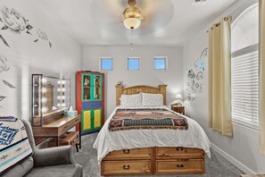 Ein Schlafzimmer mit einem hölzernen Bett, einem Frisiertisch mit Spiegel und Beleuchtung, einem farbenfrohen Kleiderschrank und einem Fenster mit Jalousien.