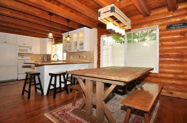Interior rústico de una cabaña con una mesa de comedor de madera, taburetes de bar, una zona de cocina y un ventanal grande.