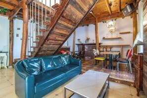 A loft-style room with exposed wooden beams, a blue leather sofa, a dining table, and a staircase.
