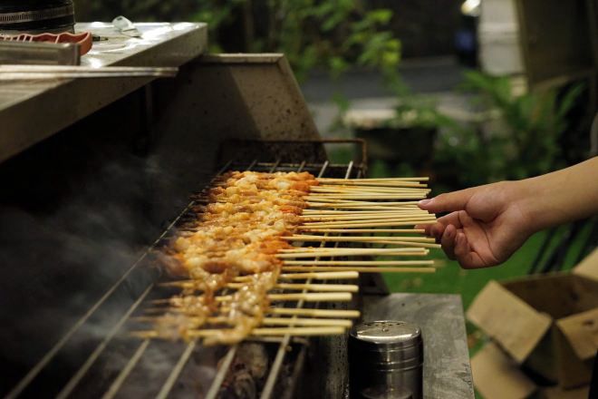 A person holding skewers over a grill with food cooking.