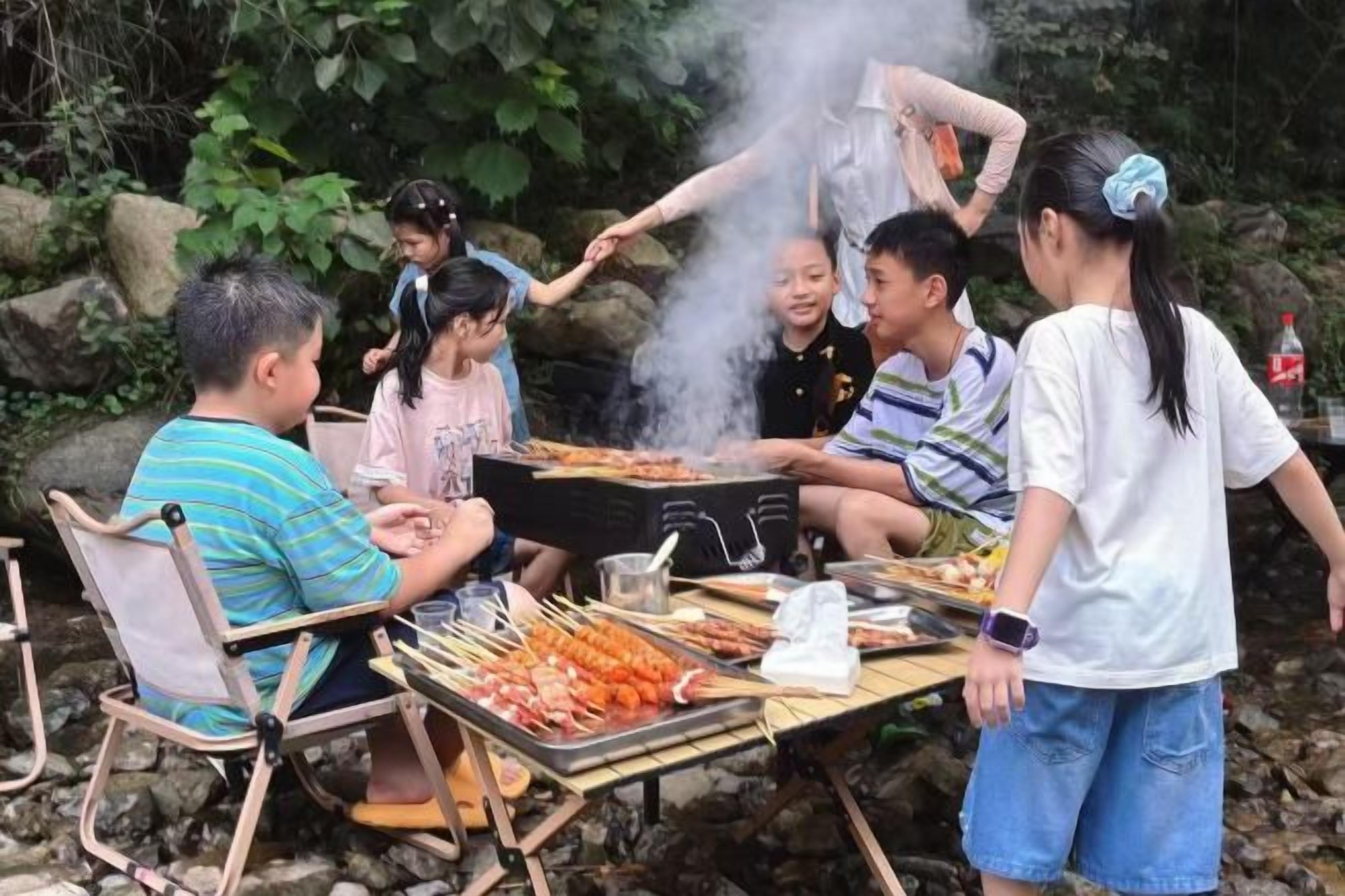 A group of people gathered around a grill with food, seated on chairs, and a table with skewers.