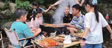 Un grupo de personas reunidas alrededor de una parrilla con comida, sentadas en sillas y una mesa con brochetas.
