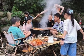 A group of people gathered around a grill with food, seated on chairs, and a table with skewers. - Yunshang Shenchu Homestay (Deqing)