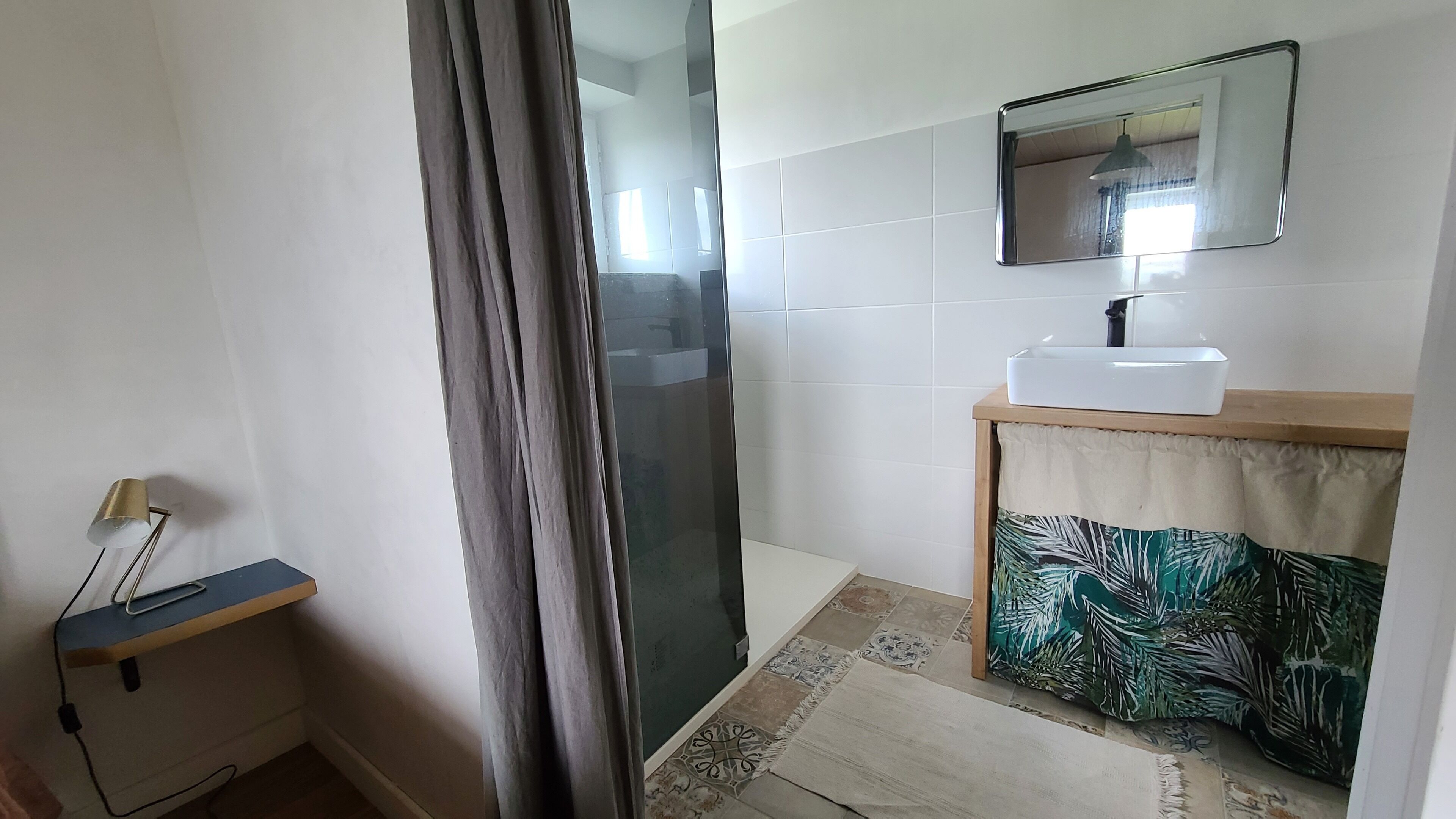 Un baño con ducha, lavamanos, espejo y un pequeño estante con lámpara.