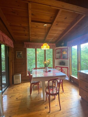 Cozy cabin interior with a dining table and four chairs, surrounded by large windows offering a forest view.