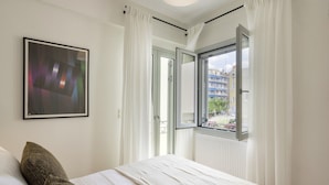Una camera da letto con un letto, tende bianche, un quadro incorniciato e una finestra vista edifici e vegetazione.