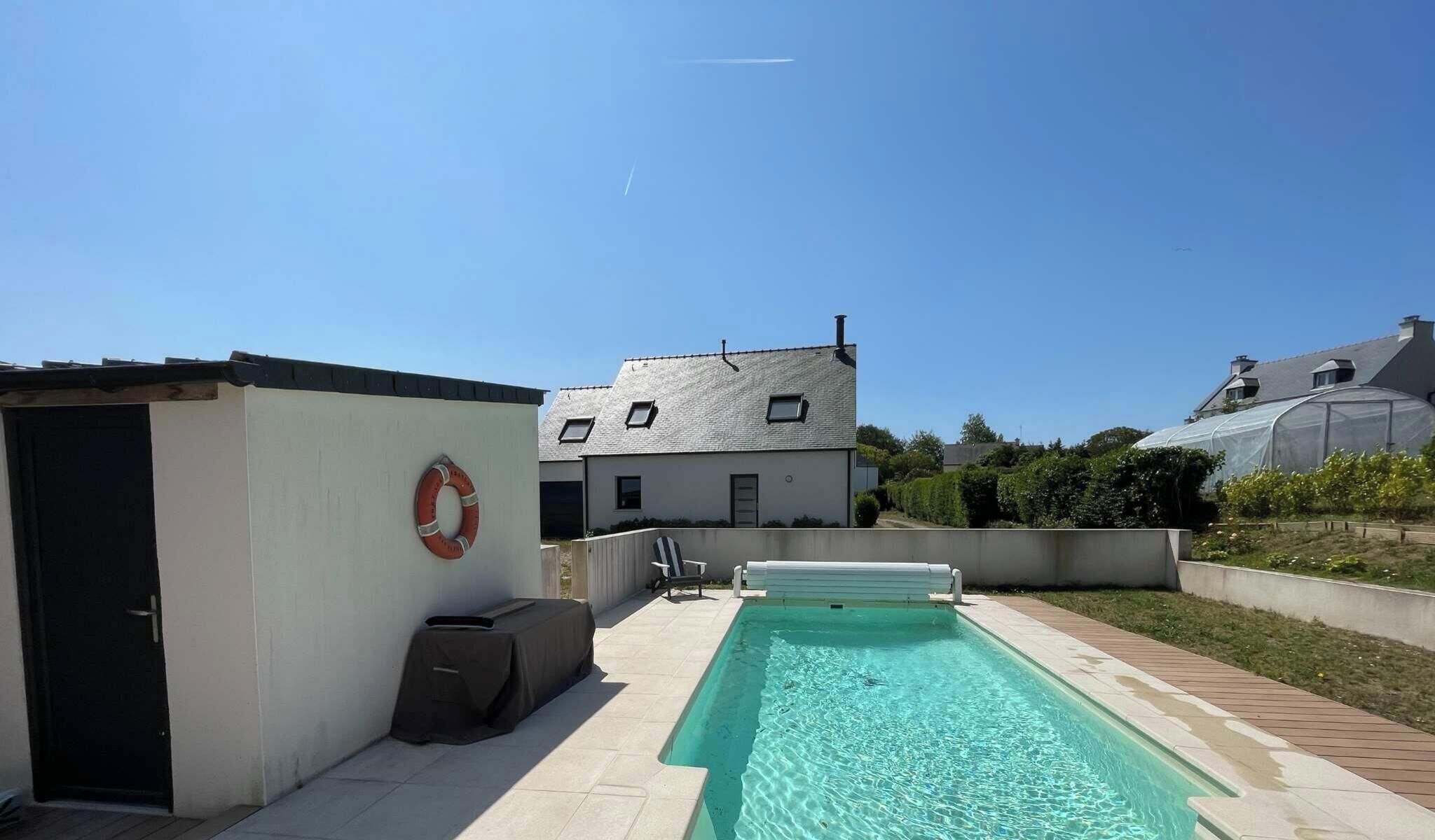Ein Schwimmbad mit Rettungsboje, ein überdachter Grillbereich und ein Haus mit geneigtem Dach.