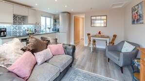 A modern living room with a grey sofa, a grey armchair, and a coffee table. There is a kitchen area with a microwave, a coffee machine, and a sink. The room has a large window and a painting on the wall. - Oswald’s Rest is in an absolutely charming location and is perfect for couples. (Bamburgh, near Seahouses)