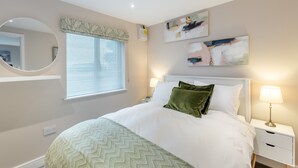 A bedroom with a bed, bedside tables, a mirror, a window with blinds, and wall art. - Oswald’s Rest is in an absolutely charming location and is perfect for couples. (Bamburgh, near Seahouses)