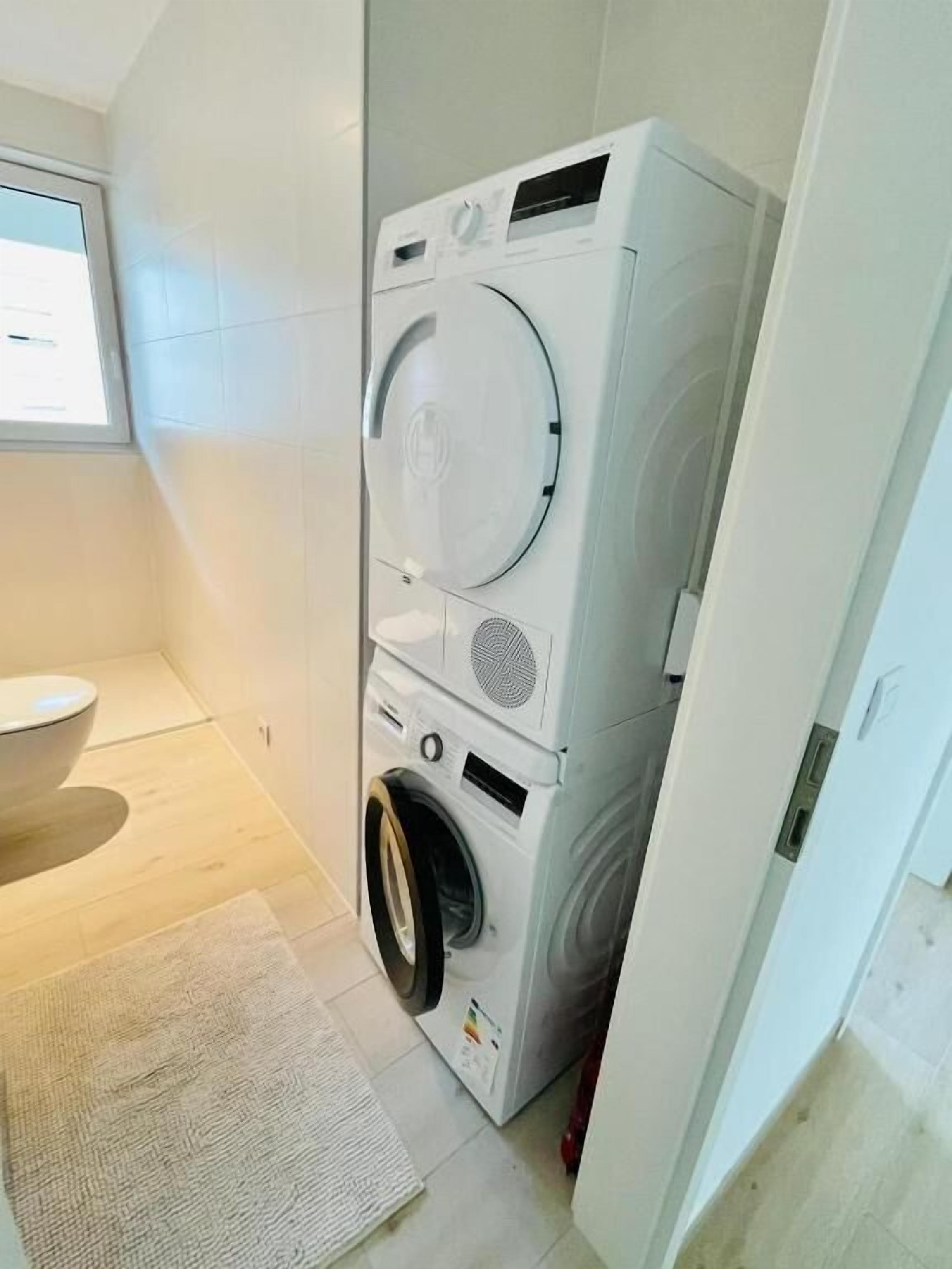 Een praktische wasruimte met een wasmachine en droger boven elkaar, een toilet en een klein raam.