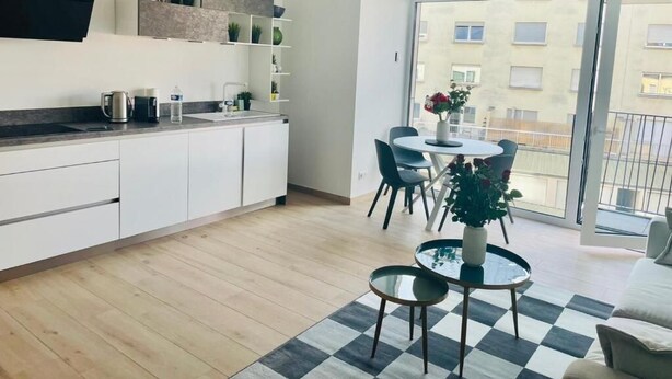 Una cocina moderna con armarios blancos, encimera negra y un comedor con mesa redonda y sillas. Hay un ventanal con vistas a edificios.