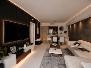 A modern living room with a flat-screen TV, a dining area, and a kitchen in the background.