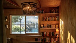 A cozy bedroom with a large window, a bookshelf, a bed with pillows, and a chandelier. - The Library at The Sea of Tranquility w/ Pool, Hot Tub & Outdoor Kitchen (Yucca Valley)