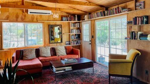 A cozy living room with a red sofa, a yellow armchair, and a coffee table. There is a bookshelf filled with books and a large window providing natural light. - The Library at The Sea of Tranquility w/ Pool, Hot Tub & Outdoor Kitchen (Yucca Valley)
