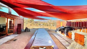 An outdoor dining area with a long table, red shade sails, and a barbecue station. - The Library at The Sea of Tranquility w/ Pool, Hot Tub & Outdoor Kitchen (Yucca Valley)