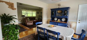 Een eetgedeelte met een tafel gedekt voor twee personen, een blauwe kast met decoratieve borden, en een uitzicht op een woonkamer met een leren bank en een raam met kantgordijnen.