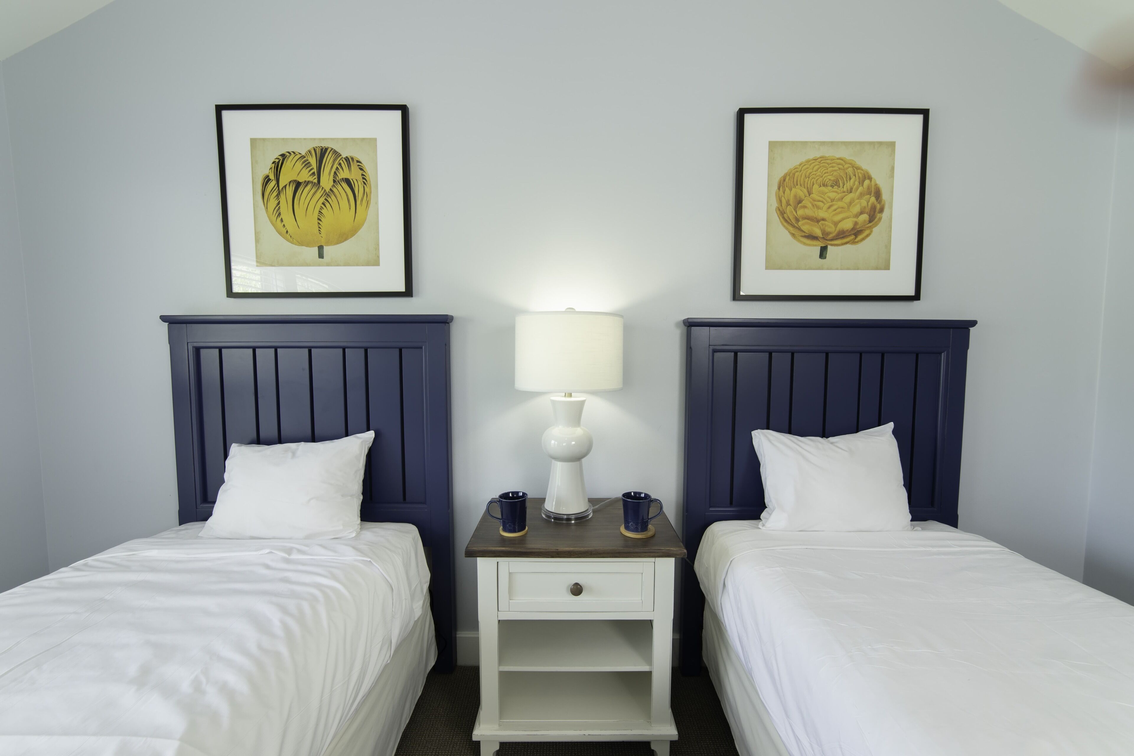 Une chambre d’hôtel avec deux lits, une table de chevet avec une lampe, deux tableaux encadrés et deux tasses.