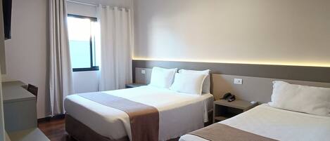 A hotel room with two beds, a desk, a chair, a window with curtains, and a ceiling light.