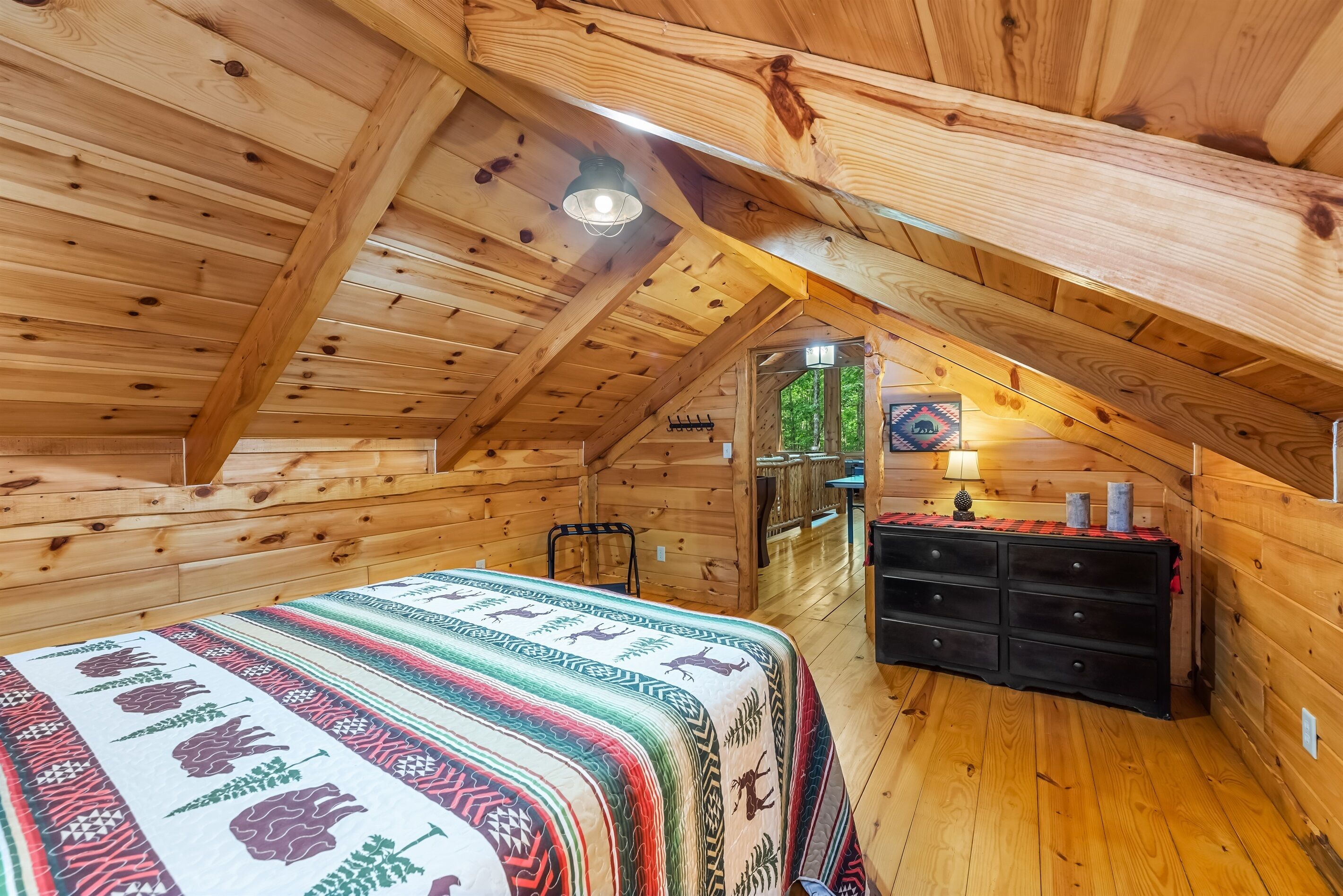 Un dormitorio acogedor en el ático, con paredes de madera y techo inclinado, que incluye una cama con una manta colorida, un aparador y una pequeña mesa con una lámpara.