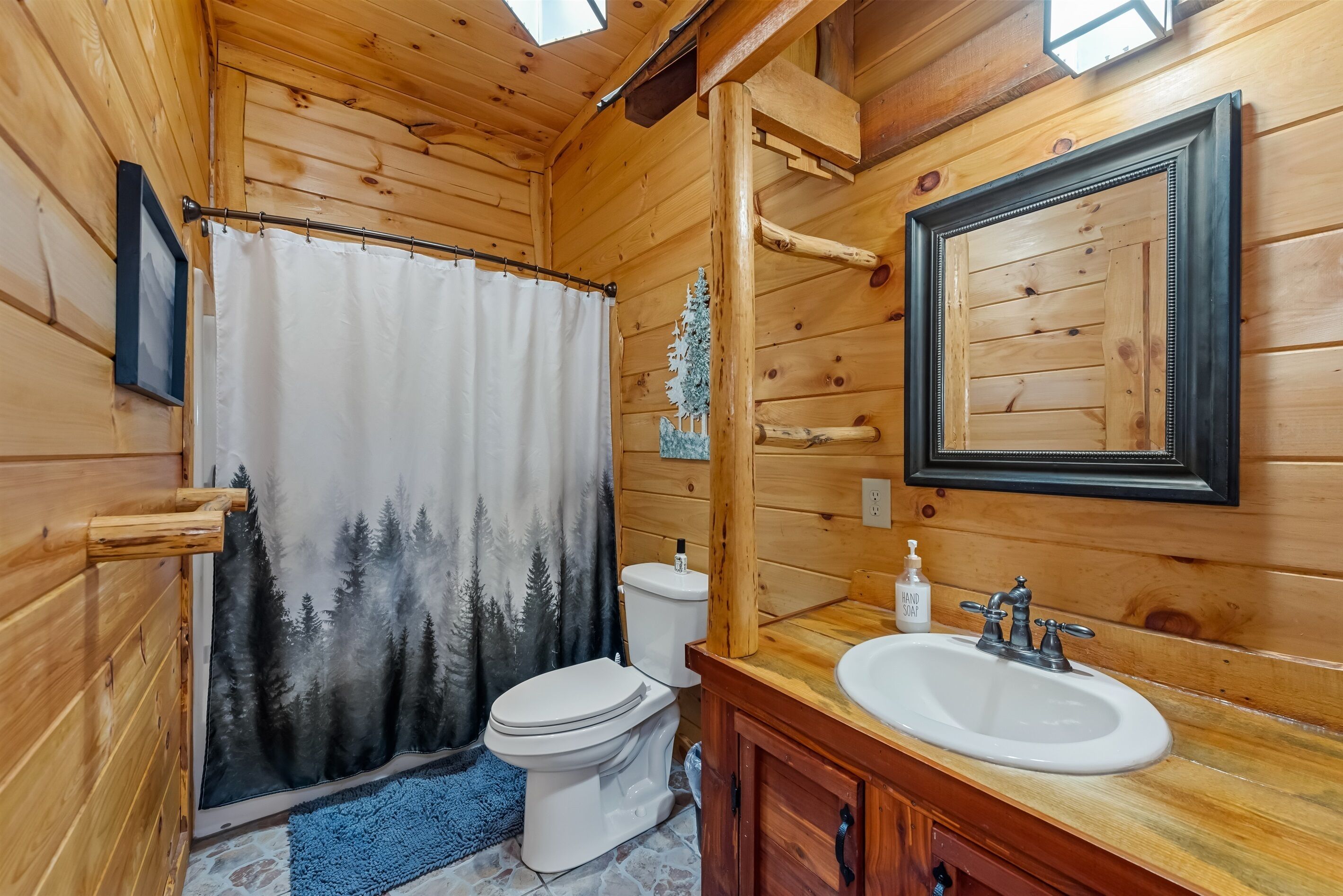 Une salle de bain rustique, aménagée dans une cabane en bois, comprenant des toilettes, un rideau de douche, un lavabo et un miroir.