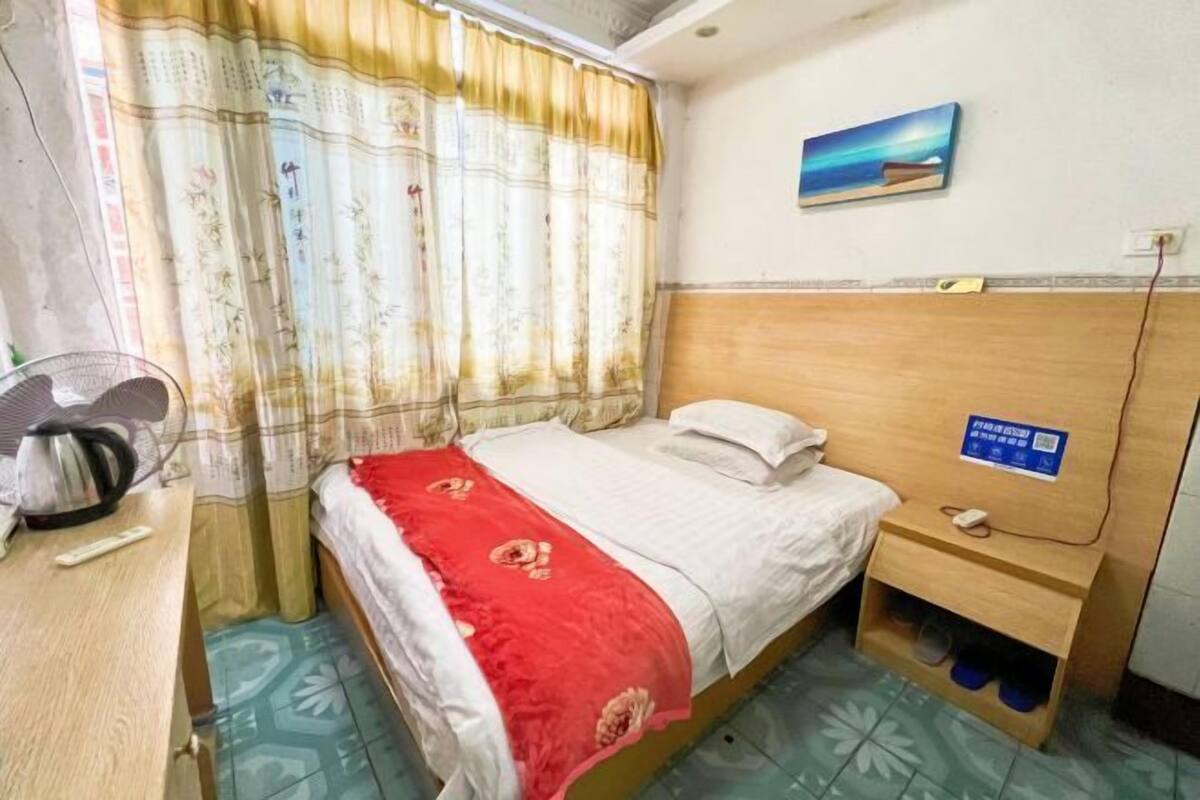 Hotellihuone, jossa on sänky, kirjoituspöytä, jossa on vedenkeitin, tuuletin, yöpöytä ja taulu seinällä.