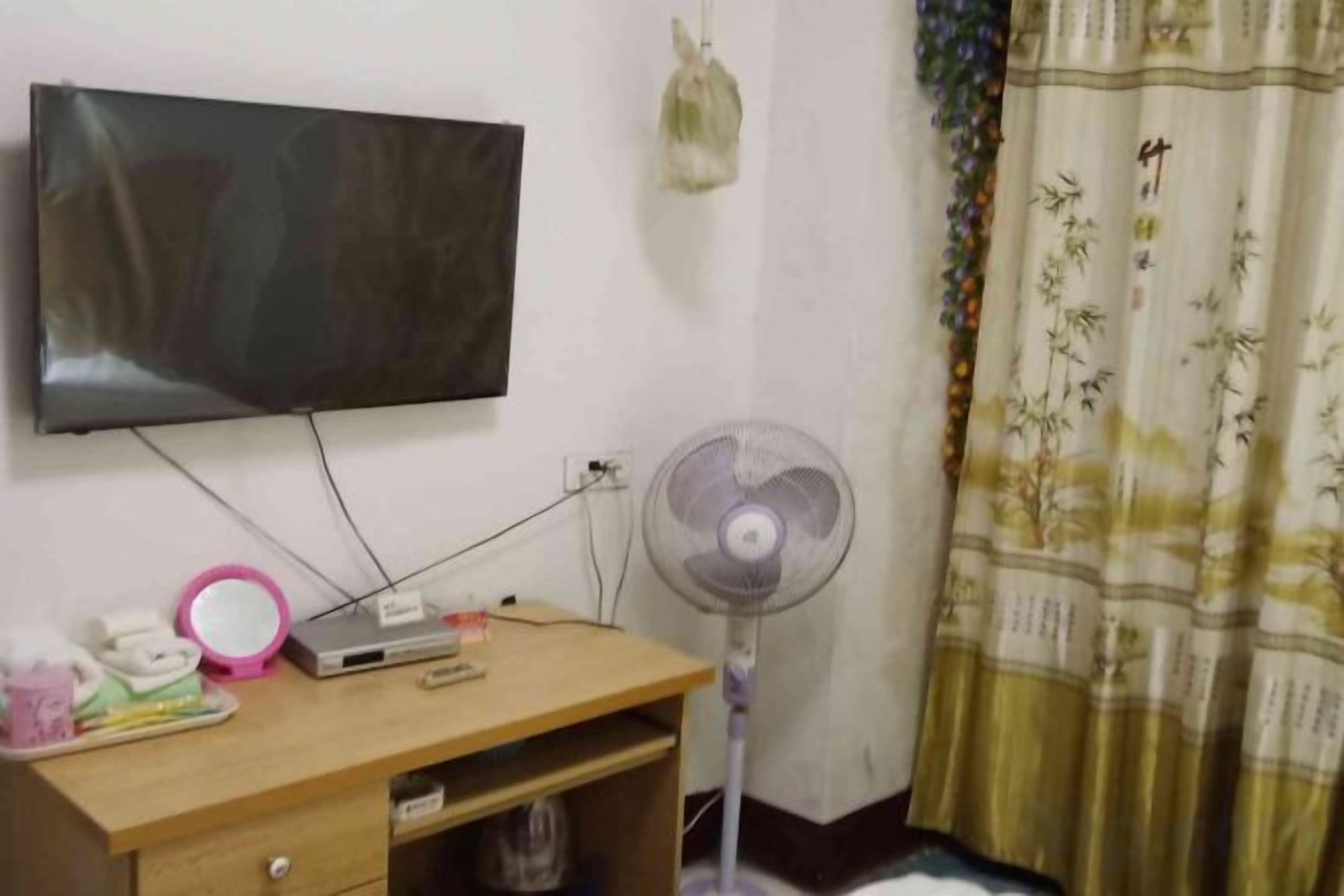Una habitación con una televisión instalada en la pared, un escritorio con un ventilador y una cortina de diseño tradicional.