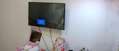 Una habitación con televisor montado, un mueble de almacenamiento rosa y una lámpara estampada.