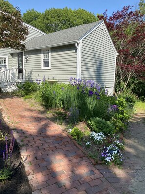 Uma casa com um caminho de tijolos que leva à porta da frente, rodeada por um jardim com diversas plantas e flores.