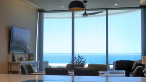 A modern living room with a large glass window, a flat-screen TV, and a dining area with a glass table. - Amazing brand new 2BD/2Bath Penthouse Condo overlooking the beach. (Puerto Peñasco)