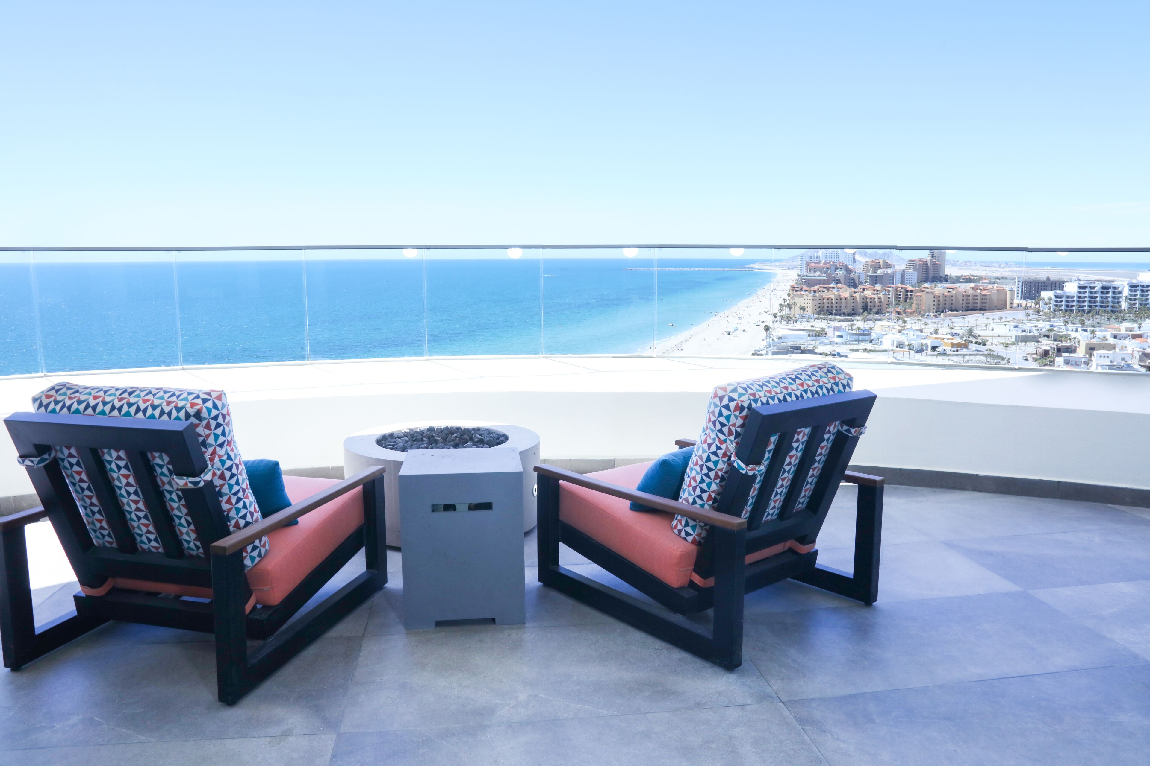 Dos sillas con cojines estampados y una pequeña mesa en un balcón con vistas a la playa y al paisaje urbano.