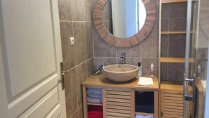 Un baño con espejo redondo, tocador de madera, lavamanos grande tipo cuenco y cabina de ducha con paredes de vidrio.