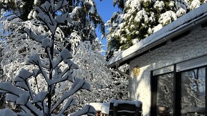 A snow-covered outdoor area with a wooden table and chairs, a stone building with large windows, and snow-laden trees.
