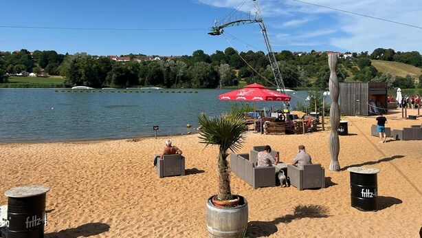 A beach with lounge chairs, a red umbrella, and a view of a lake.