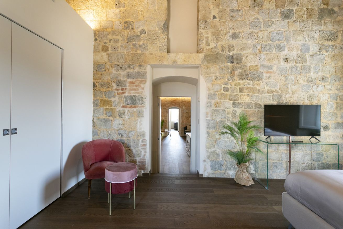 Une pièce avec un mur en pierre, un téléviseur posé sur un meuble en verre, une chaise rouge et un lit.