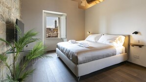 A bedroom with a bed, a window with a city view, a plant, and a wooden ceiling. - PALAZZO RINUCCINI - TORRE apartment (Siena)