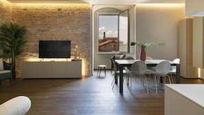 A modern living room with a brick wall, a flat-screen TV, a dining area with a table and chairs, and a large window offering a view of buildings. - PALAZZO RINUCCINI - TORRE apartment (Siena)