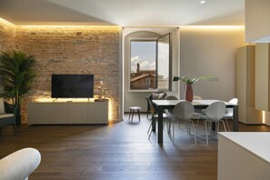 Un salon moderne avec un mur en briques, un téléviseur à écran plat, un coin repas avec une table et des chaises, et une grande fenêtre donnant sur des immeubles.