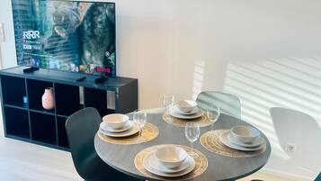 A dining area with a round table set for four, black chairs, and a green pendant light. A flat-screen TV is mounted on the wall displaying a movie.