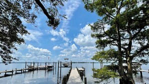 A wooden pier leading to a dock with boats, surrounded by trees and calm water under a clear sky.