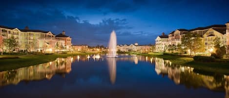A tranquil lakeside scene with a fountain, reflecting the surrounding buildings and a clear sky.