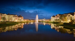 A tranquil lakeside scene with a fountain, reflecting the surrounding buildings and a clear sky. - Orlando | Compact Disney Retreat | Pool + Spa (Lake Buena Vista)