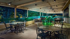 An outdoor patio area with round tables and chairs, a green railing, and a view of trees. - Orlando | Compact Disney Retreat | Pool + Spa (Lake Buena Vista)